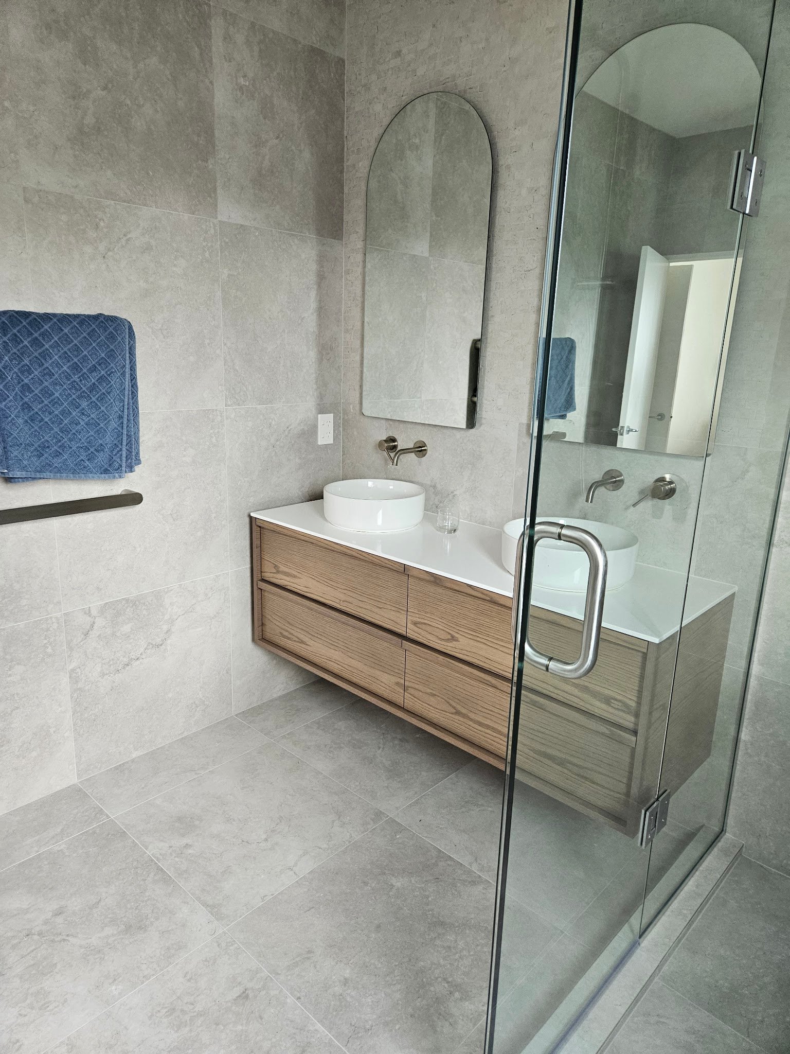 Timber double vanity with round basins and arched mirrors beside a glass shower