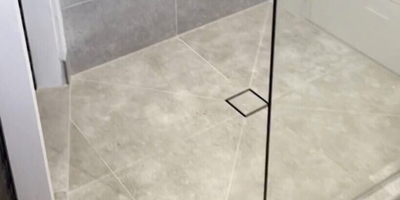Square tile-insert shower drain and frameless glass panel on pale grey floor tiles, Parnell
